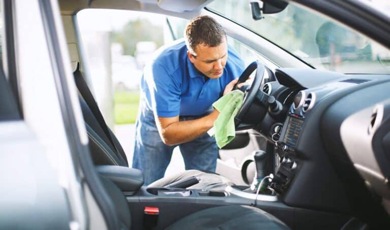 Closeup side view of mid 30's caucasian man cleaning upholstery of his car. He's sitting in driver's seat and wiping center console with soft green cloth. Both doors are ajar. This is his Sunday routine in spring and summer. Toned image.