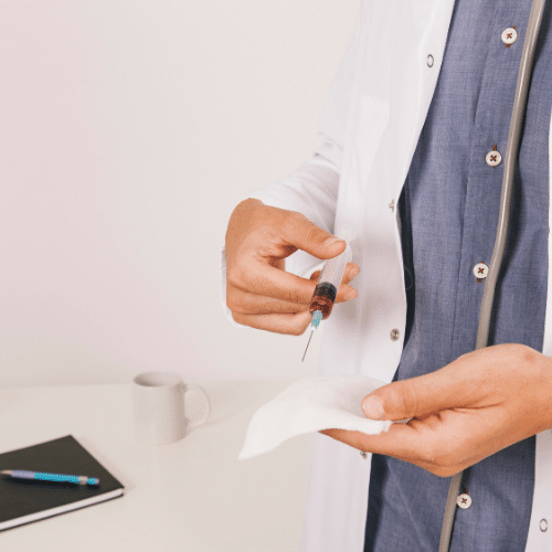 An individual in a lab coat holds a syringe filled with red liquid, alongside a sterile wipe in the opposite hand.