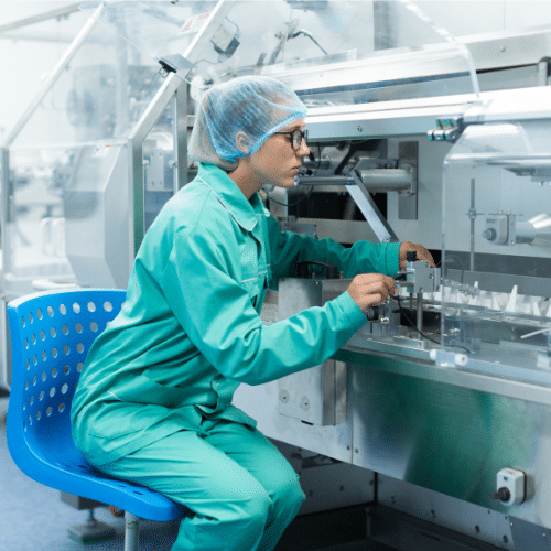 Female scientist in lab coat operating machine, wearing green overalls and blue hairnet.