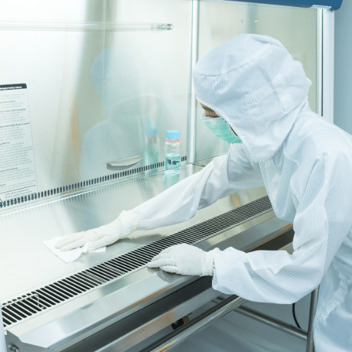 A person in a lab coat and gloves cleaning a table in a lab setting.